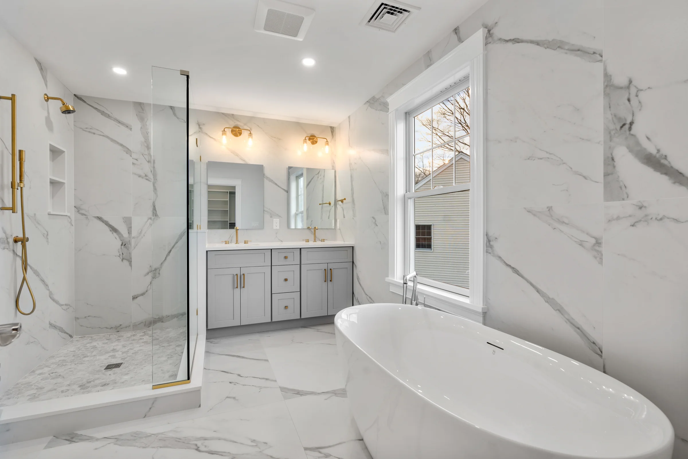 A primary bathroom in Newton with full marble walls, a freestanding tub, a glass shower, and gold fixtures: a Bay State Remodeling project