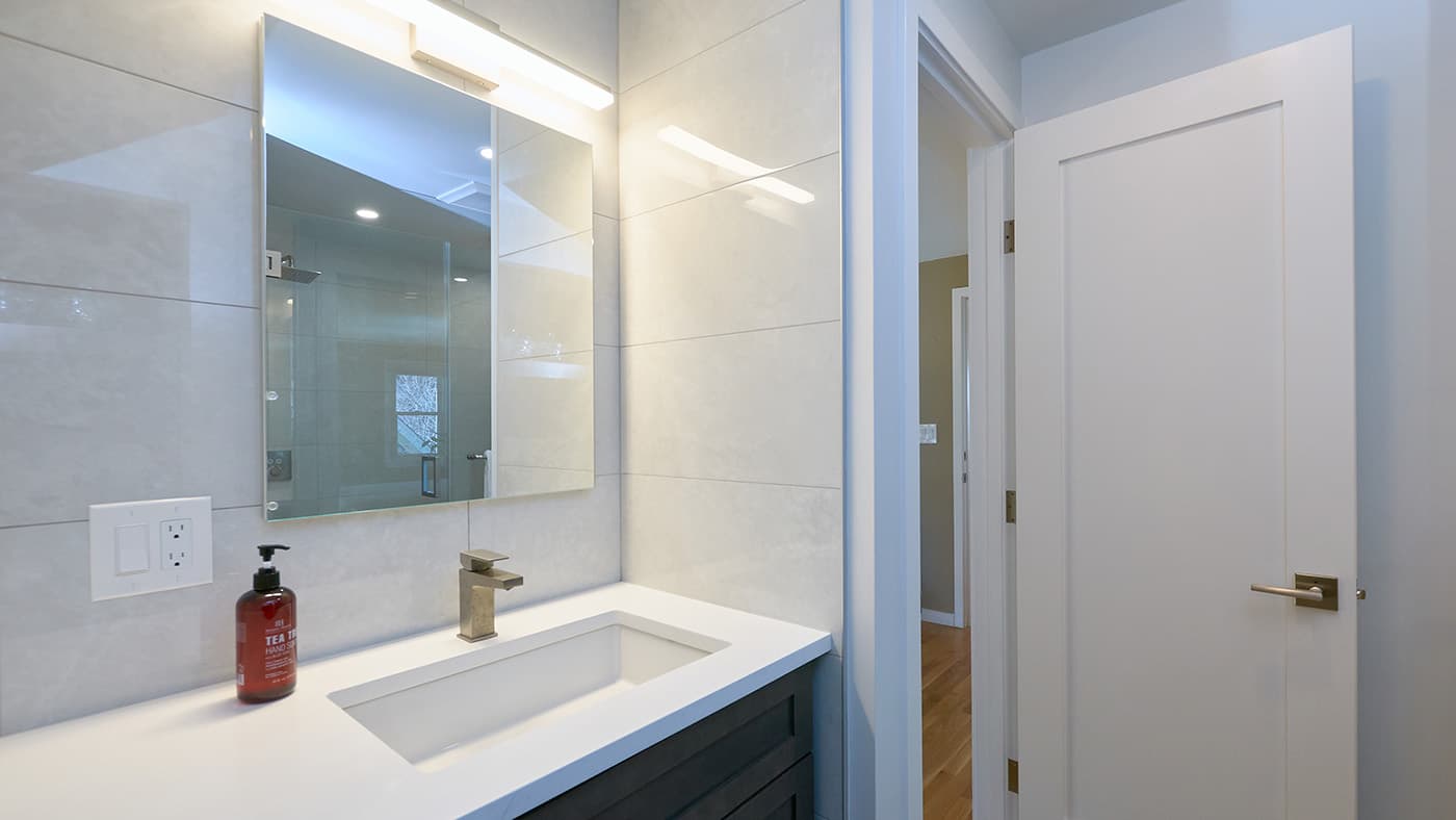 Renovated Highland Ave. Cambridge bathroom with large-format tile walls, white quartz counter, and backlit mirror