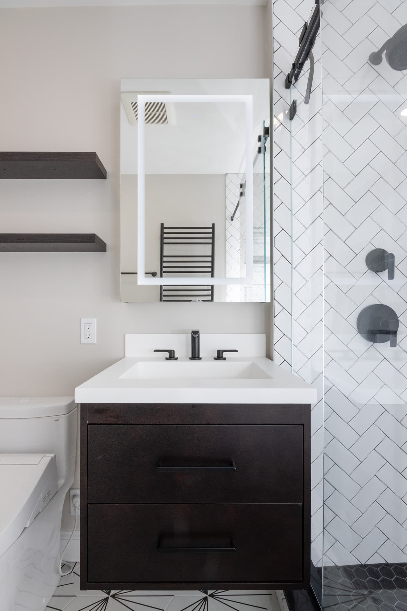 Hosmer St. bathroom after renovation in Watertown, with dark floating vanity, backlit mirror, and herringbone-tile shower.