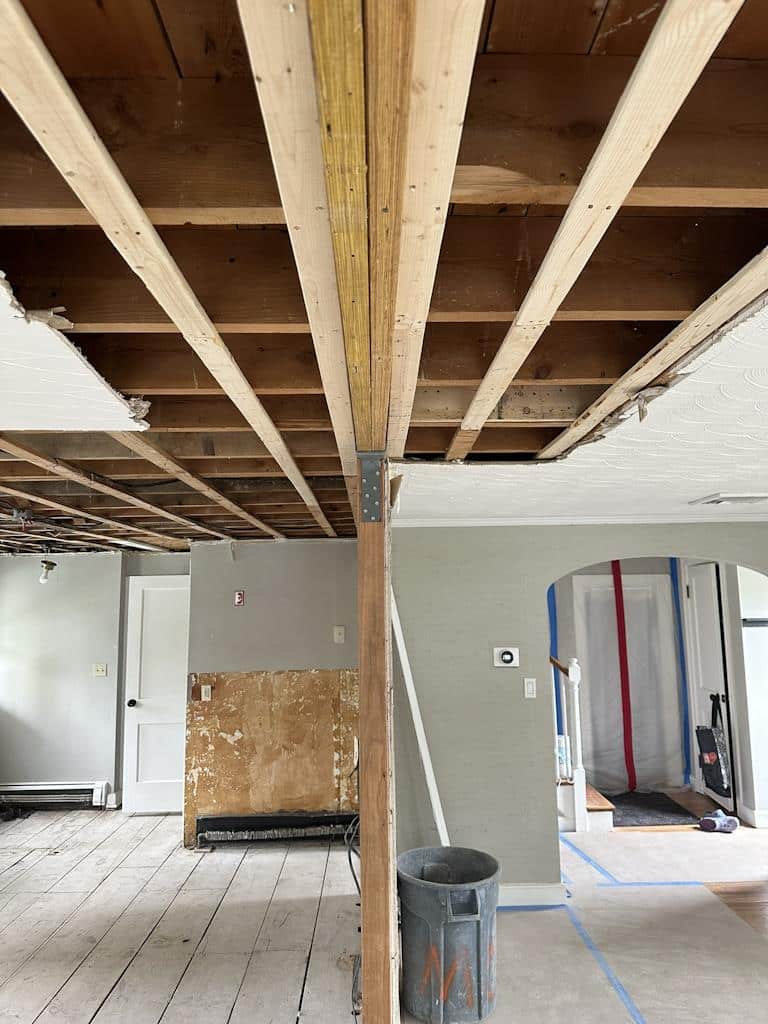 Kitchen during renovation with exposed ceiling beams, removed walls, and construction materials visible, showing structural changes in progress.