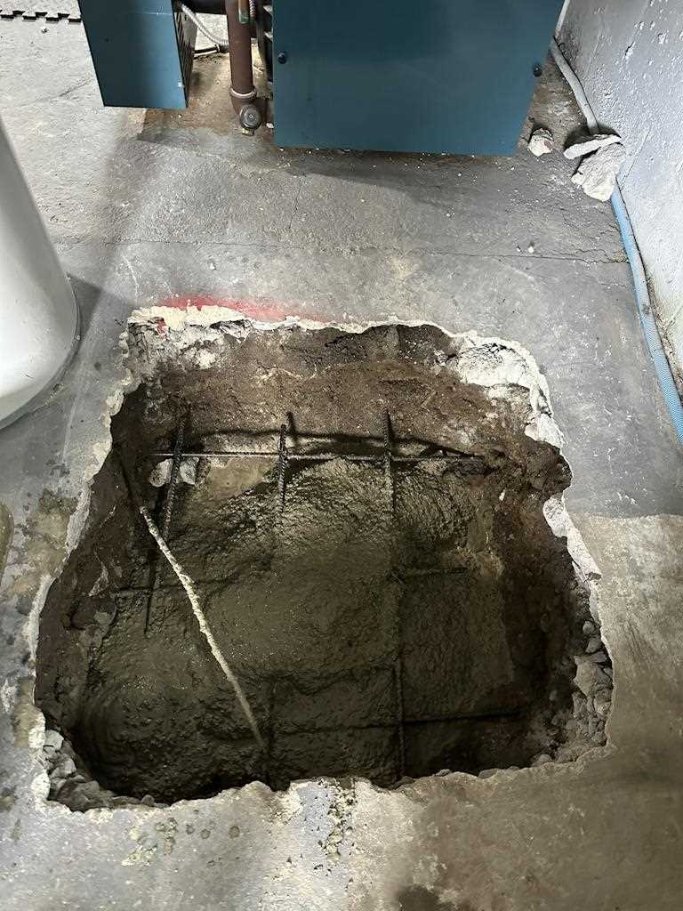 Floor section cut out with visible rebar and partially filled with concrete, adjacent to existing kitchen appliances during remodeling process.