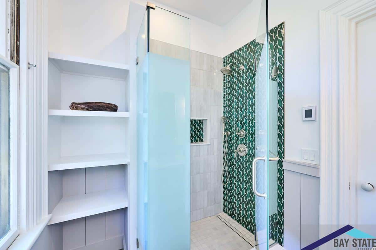 A remodeled bathroom showcasing a modern shower with green tile accents, glass doors, and built-in shelves next to the window.