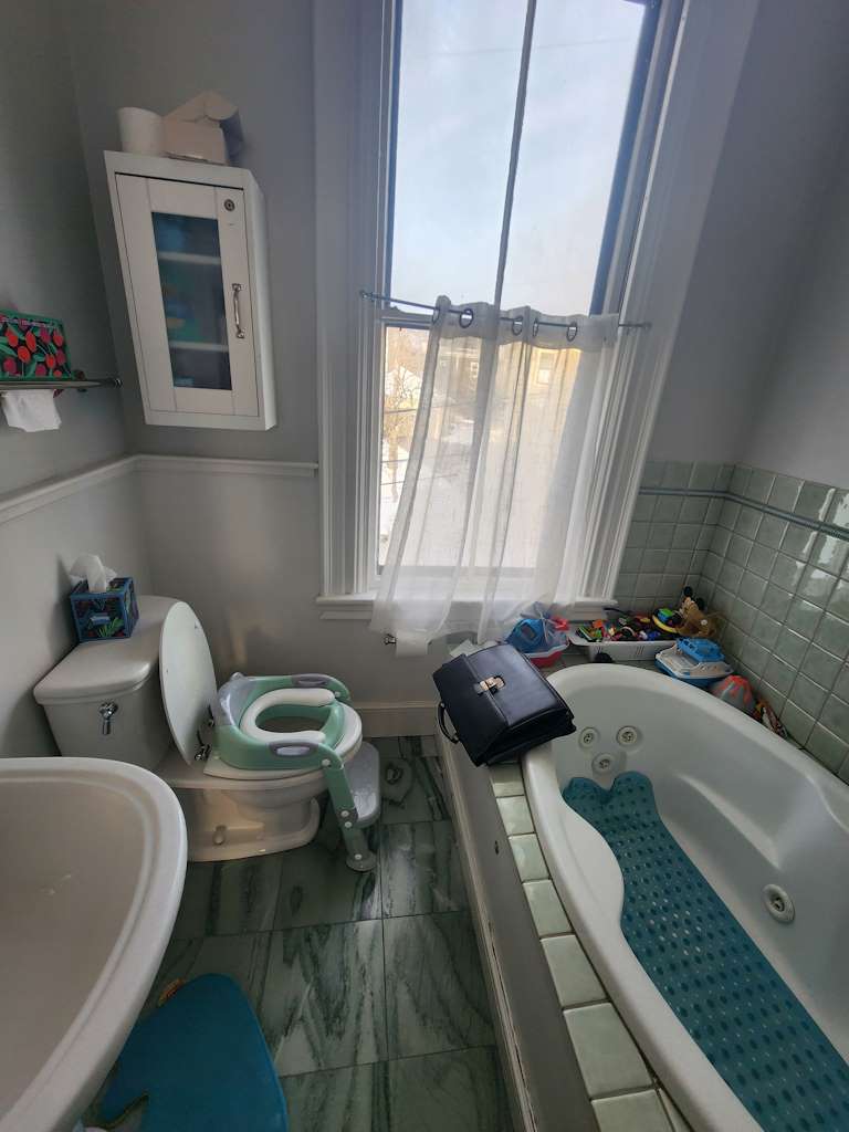 A small bathroom in need of remodeling with a toilet, sink, corner bathtub, wall cabinet, and window with sheer curtains.