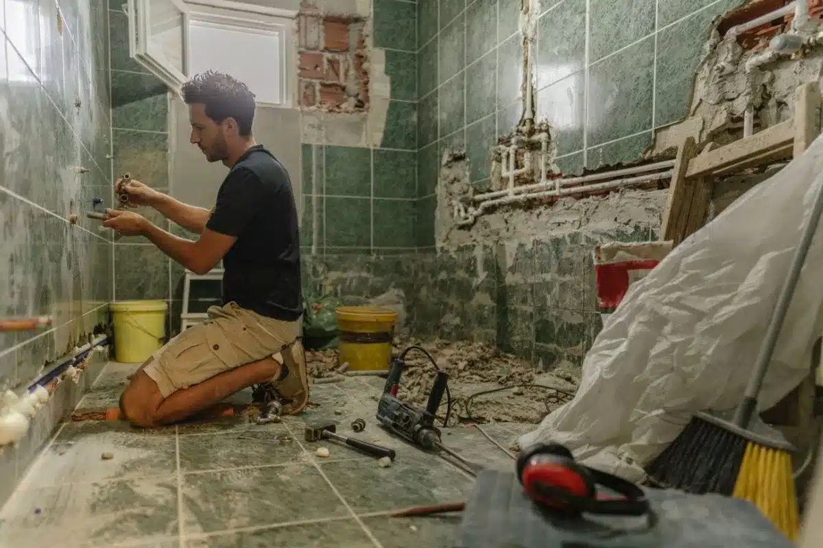 Worker repairing plumbing pipes inside a bathroom mid renovation with tools and demolition debris on the floor